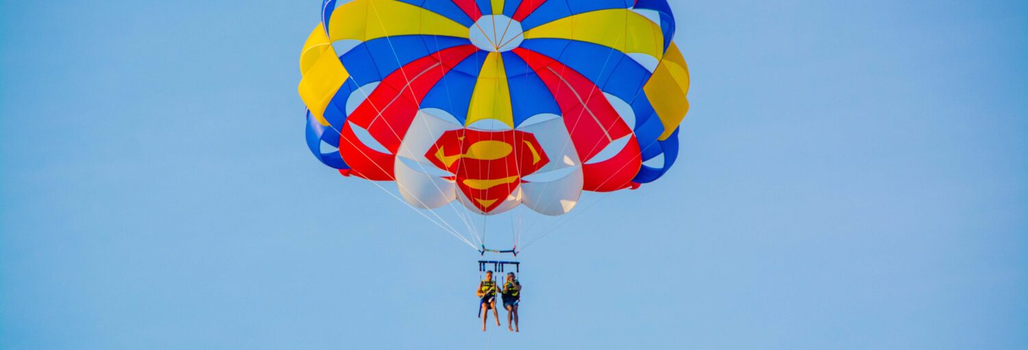Duas pessoas fazendo parasailing sob um paraquedas colorido com o logotipo do Super-Homem, em um céu azul claro. A imagem ilustra o post sobre parasailing em Barcelona.
