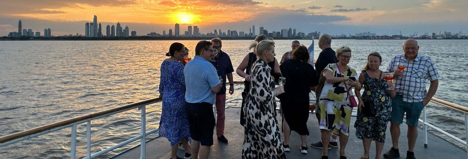 Um grupo de pessoas em pé conversando em um convés de barco ao pôr do sol, com o horizonte da cidade visível ao fundo. Imagem para ilustrar post sobre o passeio de barco ao entardecer pelo Rio da Prata.