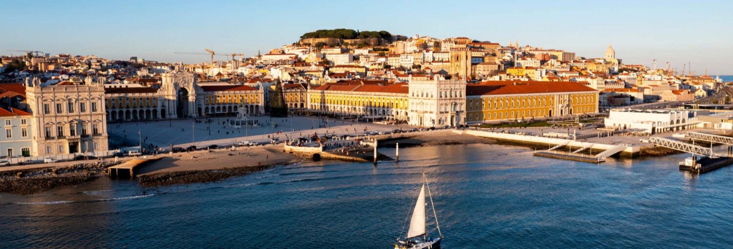 Vista aérea da Praça do Comércio de Lisboa, da zona ribeirinha e dos edifícios circundantes num dia ensolarado. Imagem para ilustrar post sobre passeio de barco com espetáculo de fado.