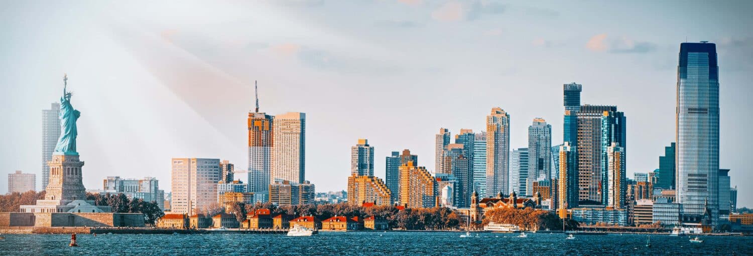 A Estátua da Liberdade e os arranha-céus se alinham na orla sob um céu parcialmente nublado, com barcos na água. A foto é capa do post Passeio de barco pela Estátua da Liberdade.