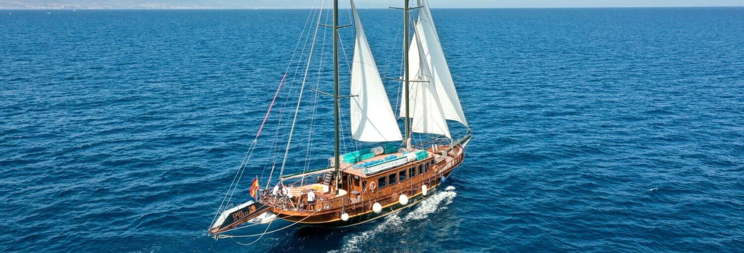 Um veleiro de madeira com dois mastros navega em um mar azul e calmo sob um céu limpo. A imagem ilustra o post sobre passeio de escuna com música em Barcelona