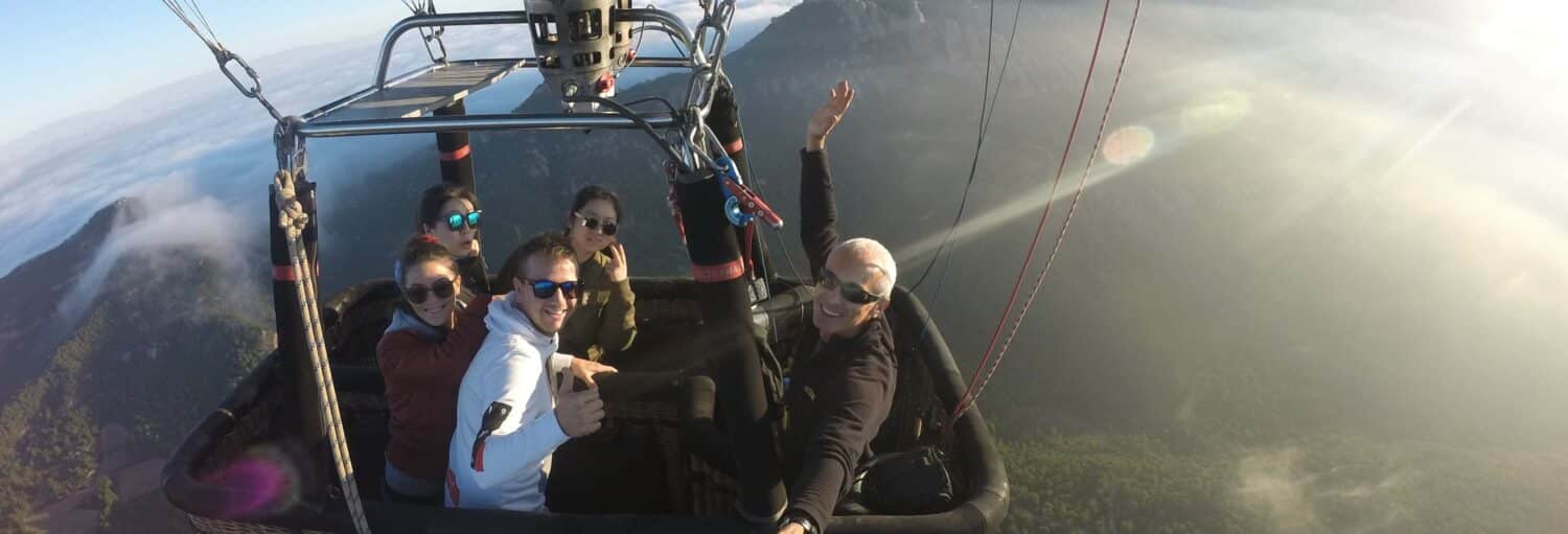 Cinco pessoas estão em uma cesta de balão de ar quente, sorrindo e acenando, com montanhas e nuvens visíveis ao fundo. A imagem ilustra o post sobre passeio de balão por Montserrat