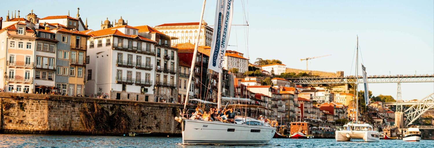 Um veleiro com passageiros desliza em um rio próximo a edifícios coloridos e uma ponte em uma cidade europeia. A foto é capa do post Passeio de catamarã ao entardecer.