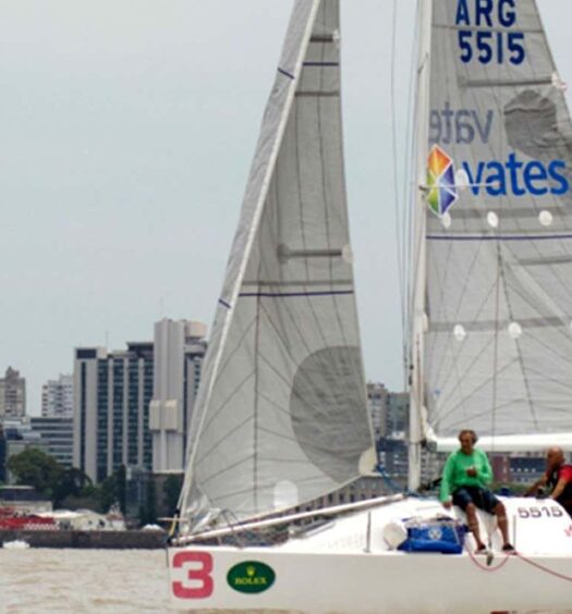 Um veleiro branco com quatro pessoas a bordo navega em um rio, com o horizonte de uma cidade ao fundo. Imagem para ilustrar post sobre passeio de veleiro pelo sul do rio da Prata.