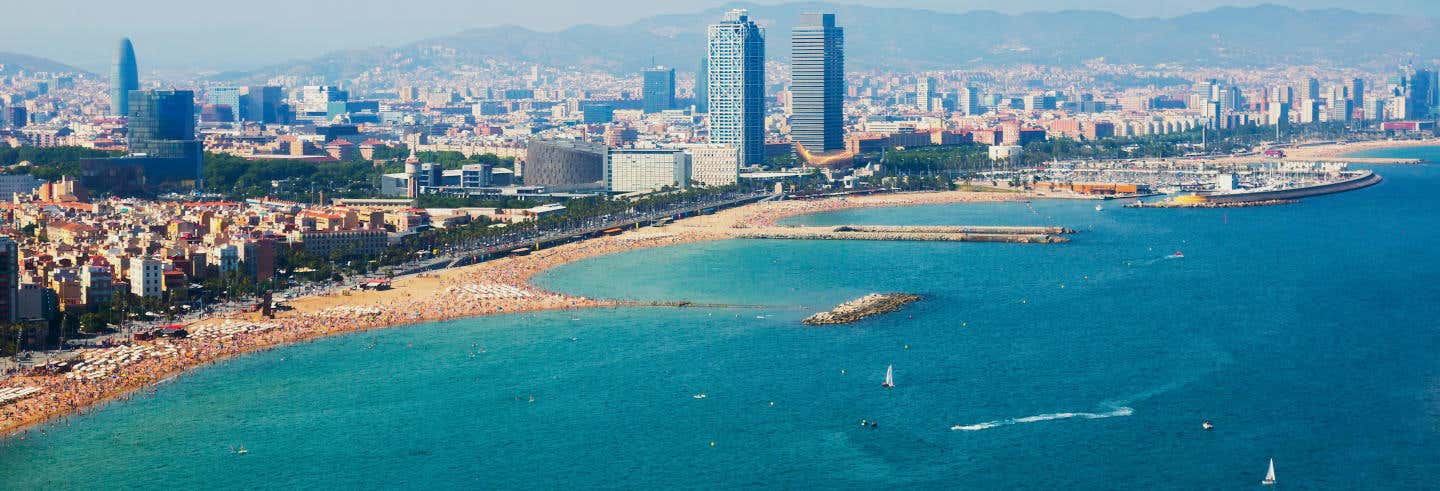 Vista aérea do horizonte de uma cidade com edifícios altos e uma praia de areia curva próxima à água azul-turquesa. A imagem ilustra o post sobre passeio de veleiro com aperitivos em Barcelona