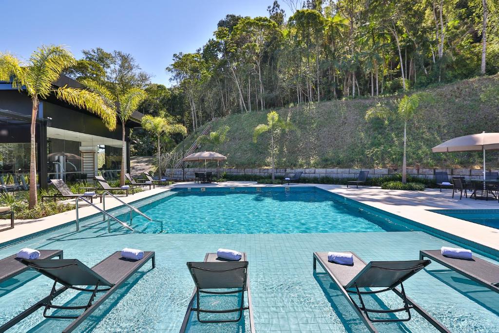 Piscina externa com espreguiçadeiras na água, cercada por árvores e guarda-sóis em um dia ensolarado, na propriedade do Quality Hotel Blumenau.