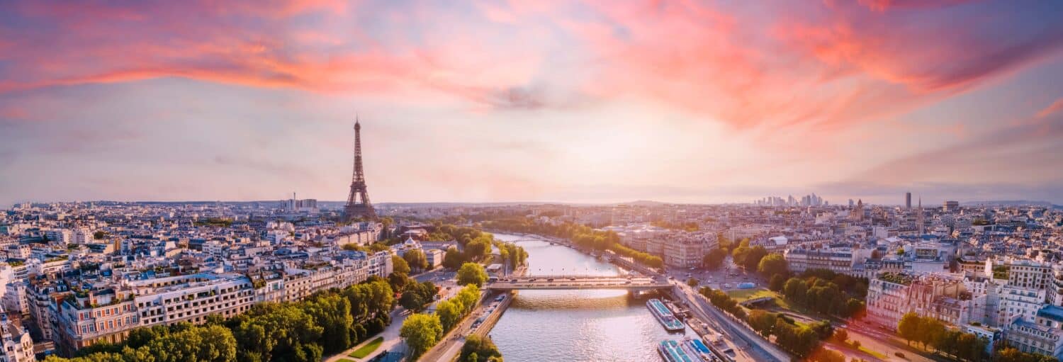 Uma vista ampla de Paris com a Torre Eiffel, o Rio Sena e os prédios da cidade sob um céu colorido de pôr do sol. A imagem ilustra o post sobre Pub Crawl. Tour por bares de Paris.