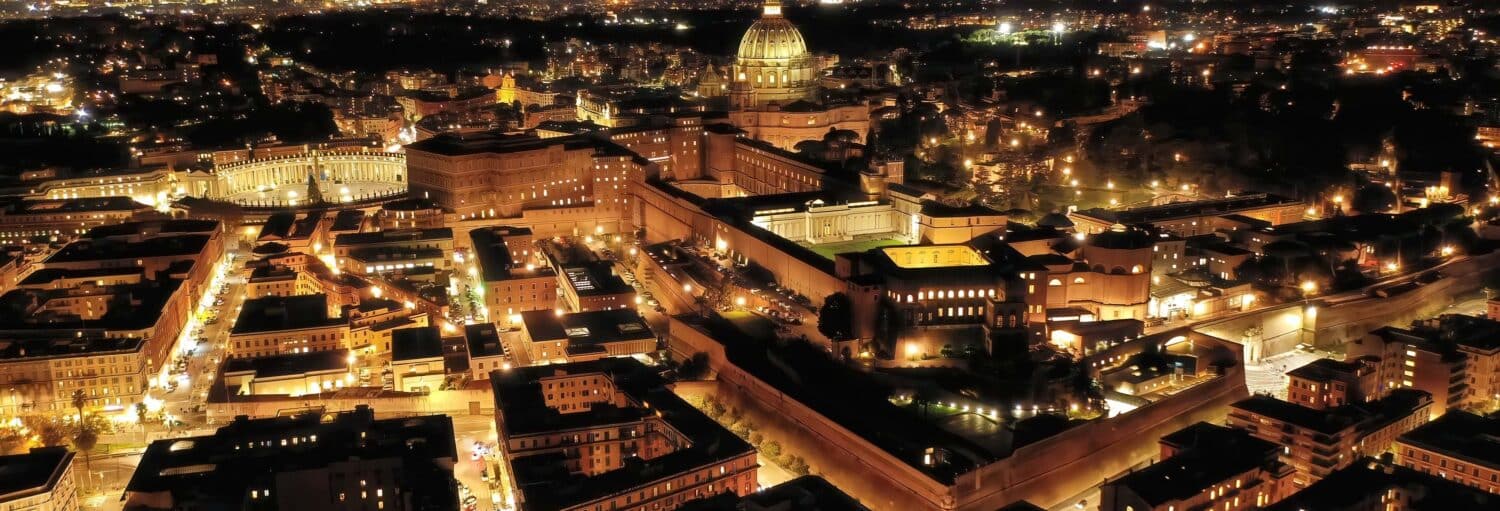 Vista aérea noturna da Cidade do Vaticano com edifícios e ruas iluminados, incluindo a Basílica de São Pedro no Pub Crawl. Tour de festa por Roma!