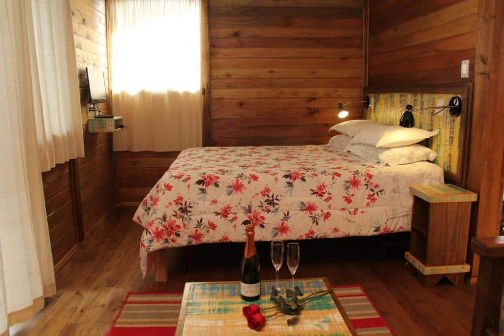 Um quarto de madeira com uma colcha floral, uma janela, uma TV e uma mesa com champanhe, copos e uma rosa, no Café e Cabanas Lenha no Fogo, para representar pousadas em Urubici.