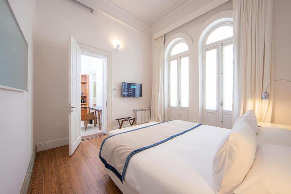 Quarto bem iluminado na Cassa Lepage Art Hotel Buenos Aires com uma cama de casal, janelas em arco, paredes brancas e uma porta aberta que leva a uma área de escritório, para representar hotéis perto da Casa Rosada em Buenos Aires.