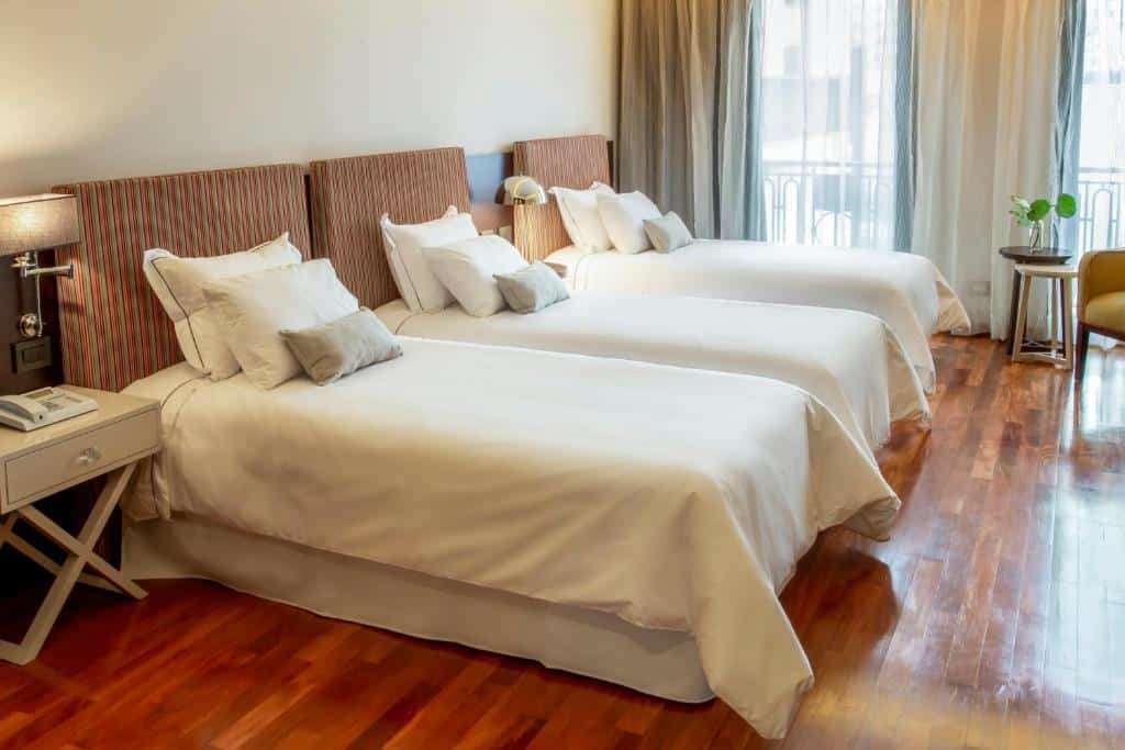 Três camas de solteiro no Esplendor by Wyndham Buenos Aires Tango com roupa de cama branca estão dispostas lado a lado em um quarto de hotel bem iluminado com piso de madeira e uma janela, para representar hotéis perto da Casa Rosada em Buenos Aires.