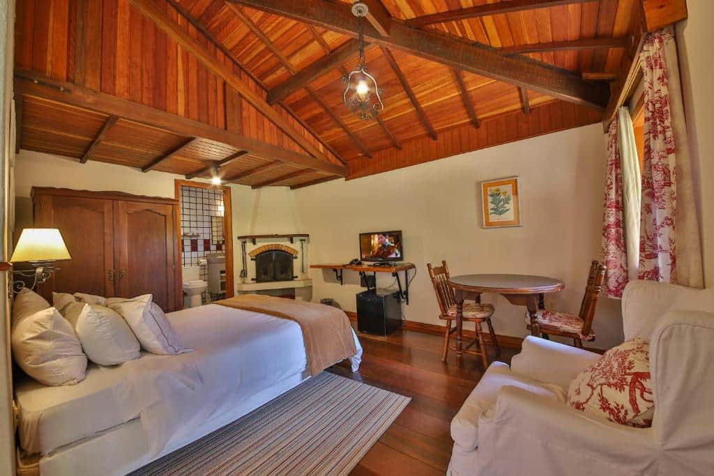 Quarto aconchegante com teto de madeira, cama de casal, poltrona, mesa redonda com cadeiras, TV e lareira, no Esquilo Hotel, para representar hotéis em Monte Verde.
