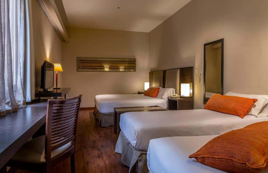 Quarto no Gran Hotel Argentino com duas camas de solteiro, travesseiros laranja, escrivaninha, cadeira, TV e iluminação aconchegante, para representar hotéis perto da Casa Rosada em Buenos Aires.