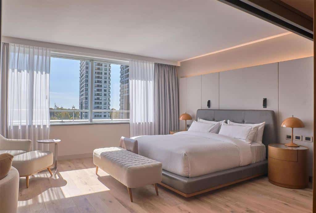 Quarto moderno no Hilton Buenos Aires com uma cama grande, duas mesas de cabeceira, um banco, poltronas e uma janela com vista para a cidade.