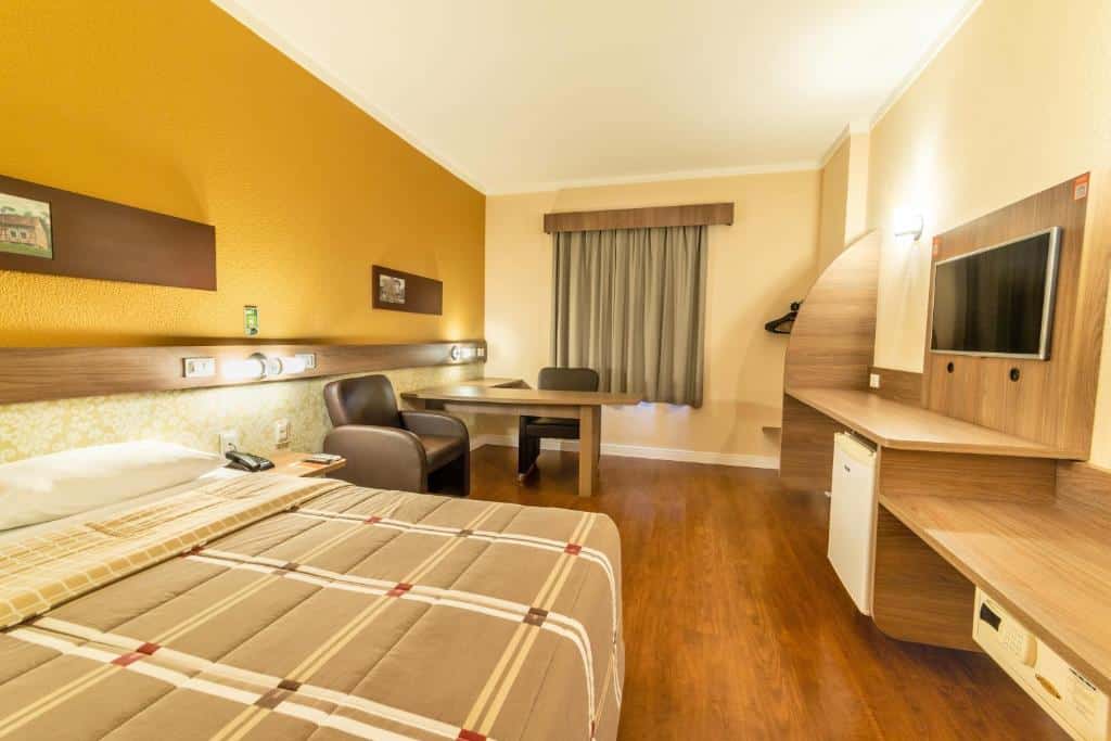 Quarto do Hotel 10 Blumenau com cama de casal, escrivaninha, cadeira, TV de tela plana, frigobar e piso de madeira, tudo em tons neutros e amarelos, para representar hotéis em Blumenau.