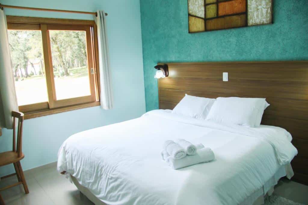 Uma cama bem arrumada com lençóis e toalhas brancas, ao lado de uma janela com uma cadeira de madeira e uma parede com detalhes em azul, no Hotel Guanxi, para representar hotéis em Monte Verde.