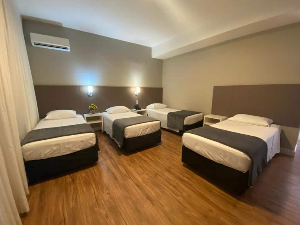 Quarto no Himmelblau Palace Hotel com quatro camas de solteiro, duas mesinhas de cabeceira, piso de madeira, luzes de parede e um aparelho de ar-condicionado, para representar hotéis em Blumenau.