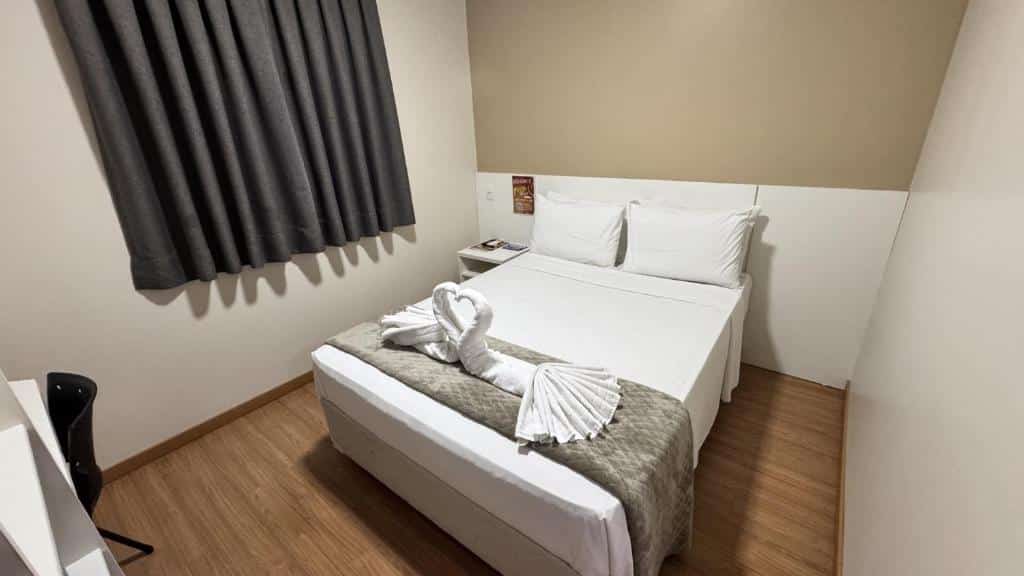 Um pequeno quarto do Hotel Zandoná com uma cama de casal, cortinas cinza, piso de madeira e toalhas dobradas em forma de cisne sobre a cama, para representar hotéis em Blumenau.