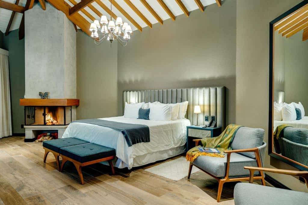 Quarto moderno com uma cama grande, lareira, poltrona, lustre, piso de madeira e um grande espelho na parede, na Pousada Spa Mirante da Colyna, para representar pousadas em Monte Verde.