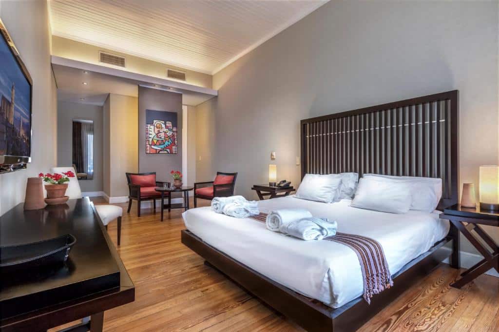 Quarto no Patios de San Telmo com uma cama grande, duas cadeiras, uma escrivaninha, uma TV montada e obras de arte na parede sobre um piso de madeira, para representar melhores hotéis em Buenos Aires.