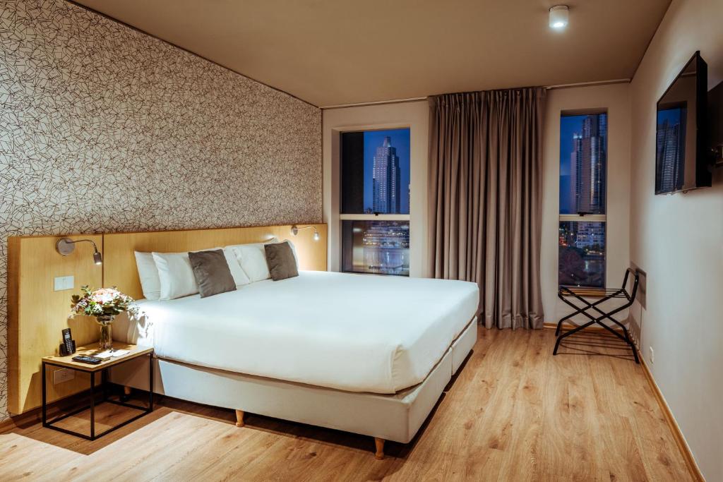 Quarto no Pleno Madero com uma cama grande, mesa de apoio, TV de tela plana e vista da cidade através de duas janelas à noite, para representar hotéis peto da Casa Rosada em Buenos Aires.