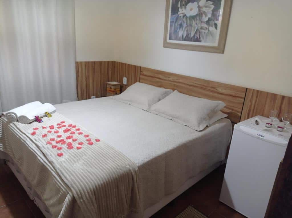 Uma cama bem arrumada com lençóis brancos, um pequeno frigobar, toalhas e pétalas em um cobertor em um quarto com decoração simples, na Pousada Alpenblumen, para representar pousadas em Monte Verde.
