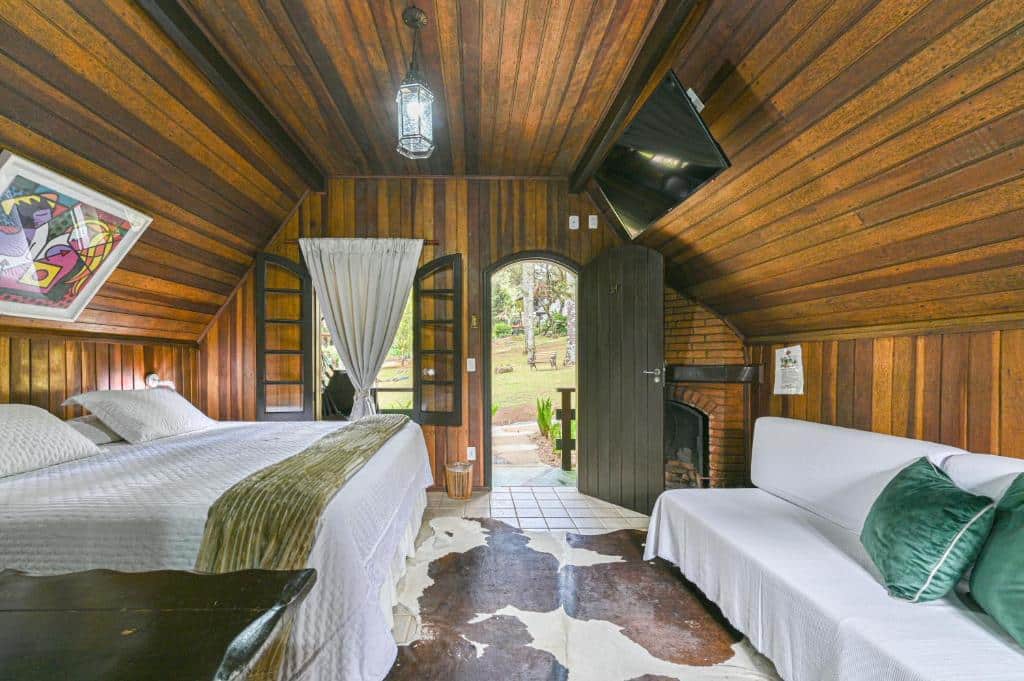 Um quarto aconchegante em cabine de madeira com cama de casal, sofá branco, porta aberta em arco, tapete de pele de vaca e vista para um jardim externo, na Pousada Cantos e Contos, para representar pousadas em Monte Verde.