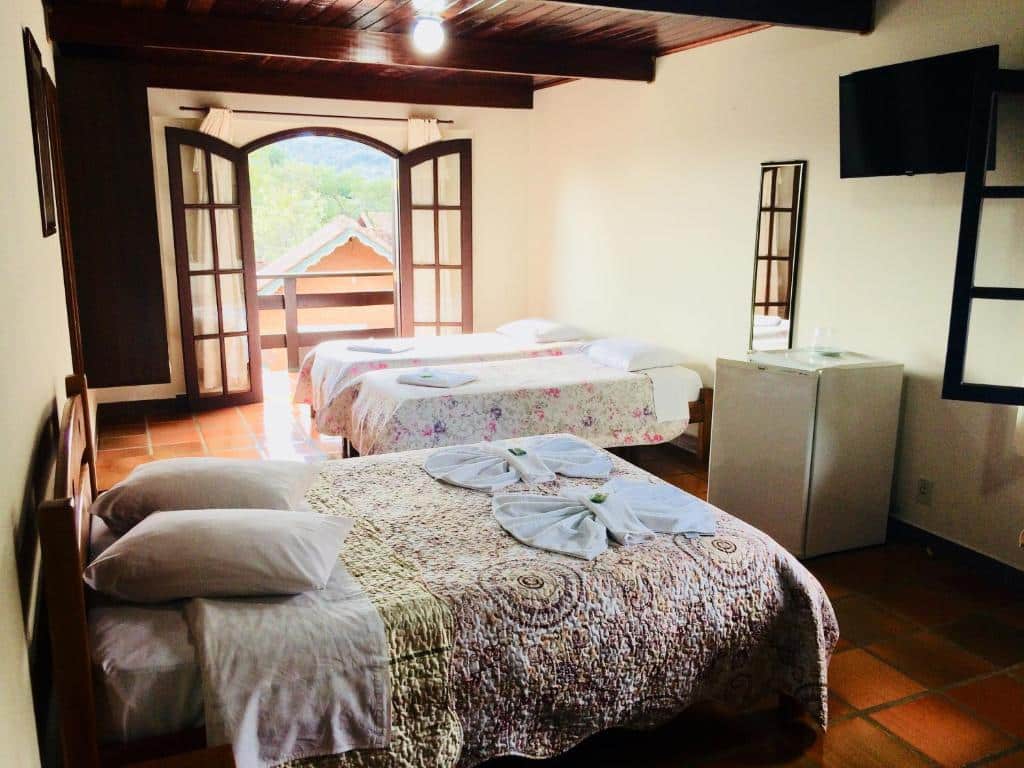 Quarto com duas camas, um pequeno frigobar, TV na parede, portas abertas para uma varanda e entrada de luz do dia, na Pousada Dona Ana, para representar pousadas em Monte Verde.