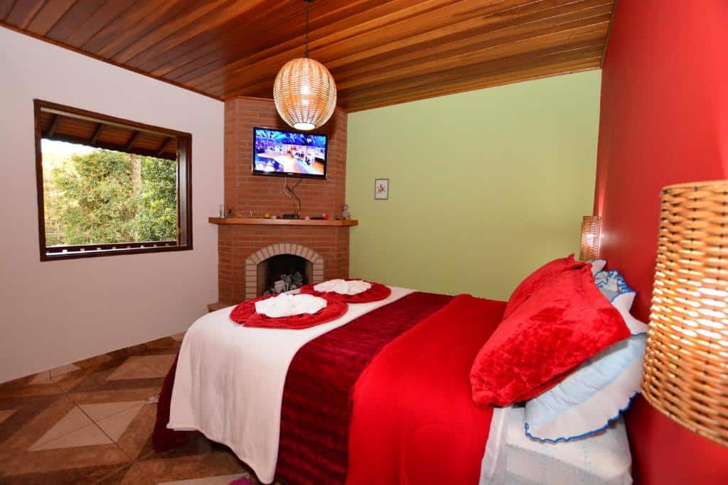 Um quarto aconchegante com uma cama de casal, roupa de cama vermelha, uma lareira de tijolos, uma TV e uma janela com vista para as árvores, na Pousada Florada da Serra, para representar pousadas em Monte Verde.