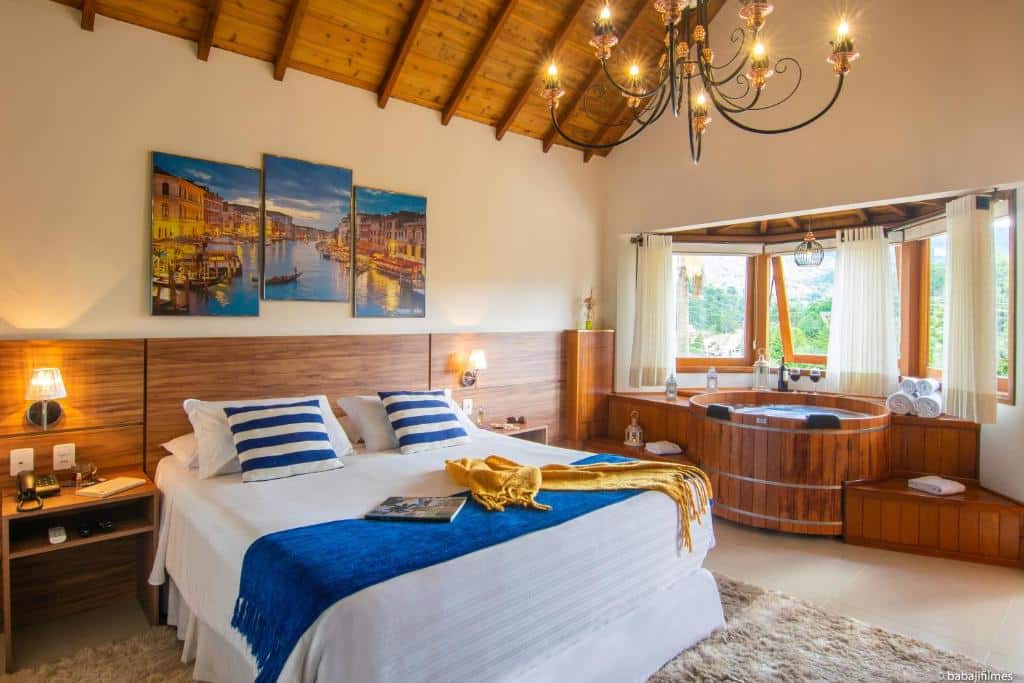 Um quarto de hotel aconchegante com uma cama de casal, detalhes em azul, obras de arte na parede e uma banheira de hidromassagem de madeira ao lado de uma grande janela.