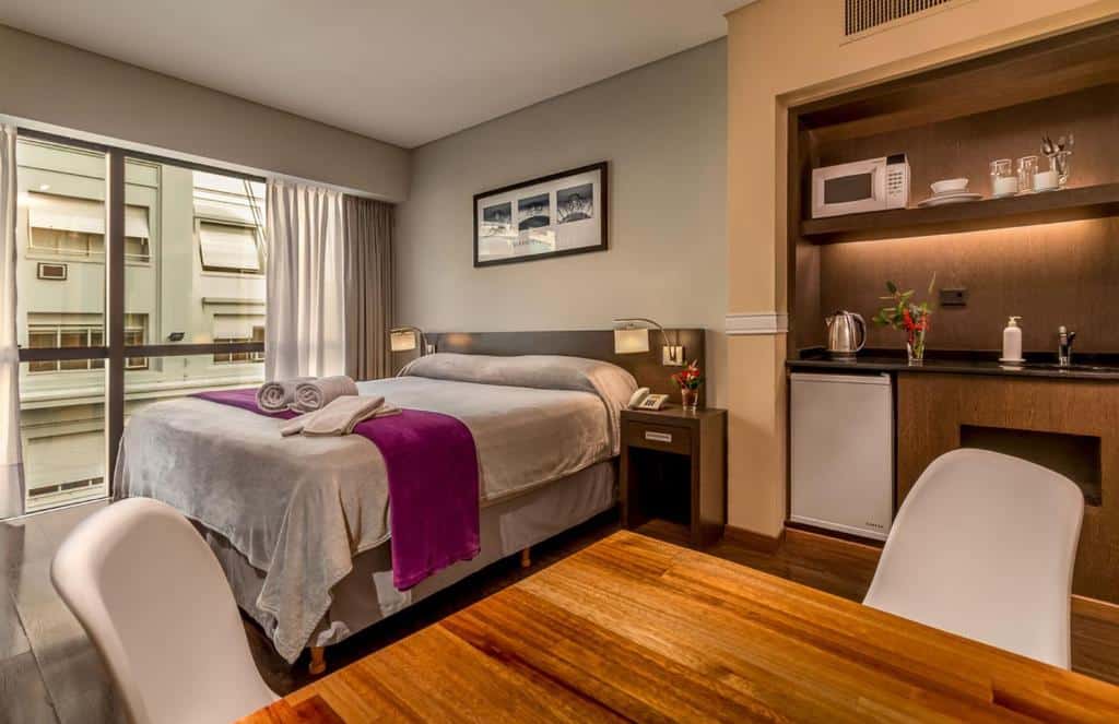 Um quarto moderno no Up Viamonte Hotel com uma cama queen, cozinha compacta, pequena mesa de jantar e uma janela com vista para os edifícios próximos, para representar onde ficar em Buenos Aires.