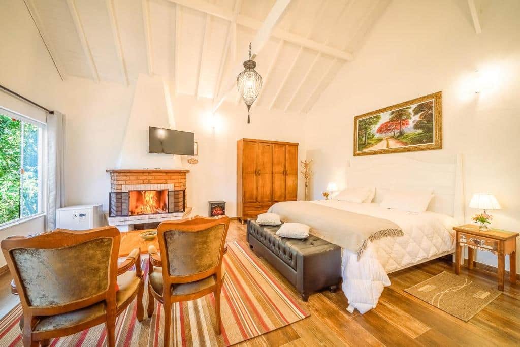 Quarto bem iluminado com lareira, cama, poltronas, TV, móveis de madeira e uma grande janela que permite a entrada de luz natural, no VELINN Pousada Jardim da Mantiqueira, para representar pousadas em Monte Verde.