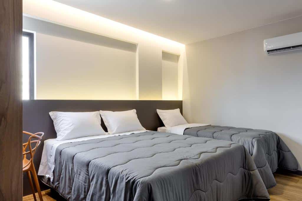 Um quarto no Villano Hotel com duas camas, roupa de cama cinza, travesseiros brancos, uma cadeira de madeira e um aparelho de ar-condicionado na parede, para representar hotéis em Blumenau.
