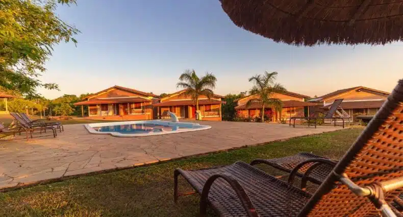 Espreguiçadeiras e uma piscina com várias casas de telhado vermelho e palmeiras ao fundo sob um céu claro.