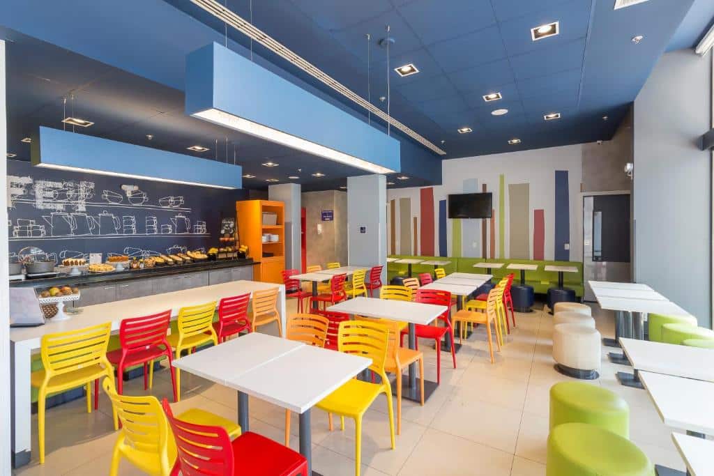 Uma cafeteria moderna com cadeiras coloridas, mesas brancas, um bufê de café da manhã e uma parede com quadro-negro atrás do balcão, na propriedade do Ibis Budget Blumenau.