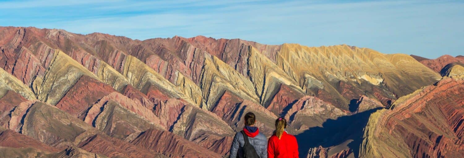 Duas pessoas estão diante de colinas multicoloridas com listras em ziguezague sob um céu azul na cordilheira Hornocal. Imagem para ilustrar post sobre Buenos Aires, Salta, Jujuy e Tucumán em 9 dias.