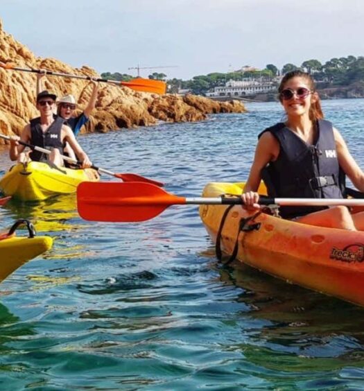 Três pessoas usando coletes salva-vidas remam em caiaques amarelos e laranja perto de penhascos rochosos em águas azuis claras. A imagem ilustra o post sobre trilha, caiaque e snorkel na Costa Brava em Barcelona.