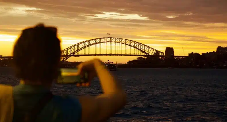 Pessoa tirando uma foto da Sydney Harbour Bridge ao pôr do sol, com água e o horizonte da cidade ao fundo. A foto é capa do post de eSIM Sydney.