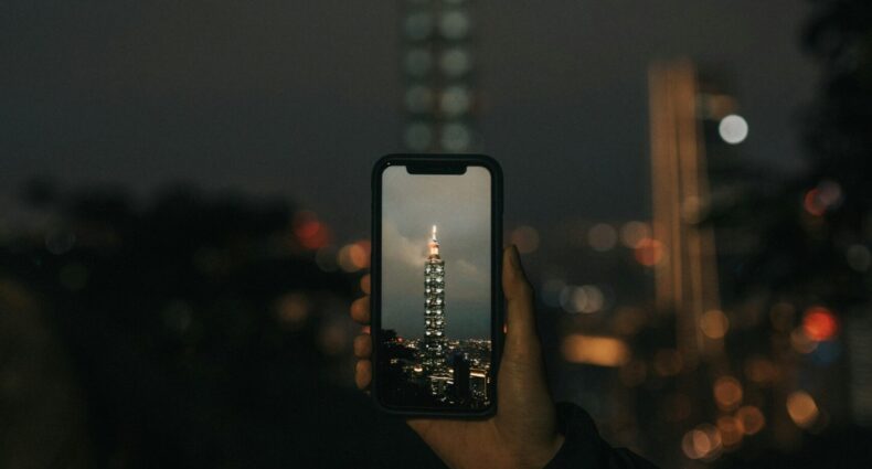 Uma mão segura um telefone capturando um arranha-céu do Taipei 101 iluminado à noite, com o edifício real desfocado ao fundo.