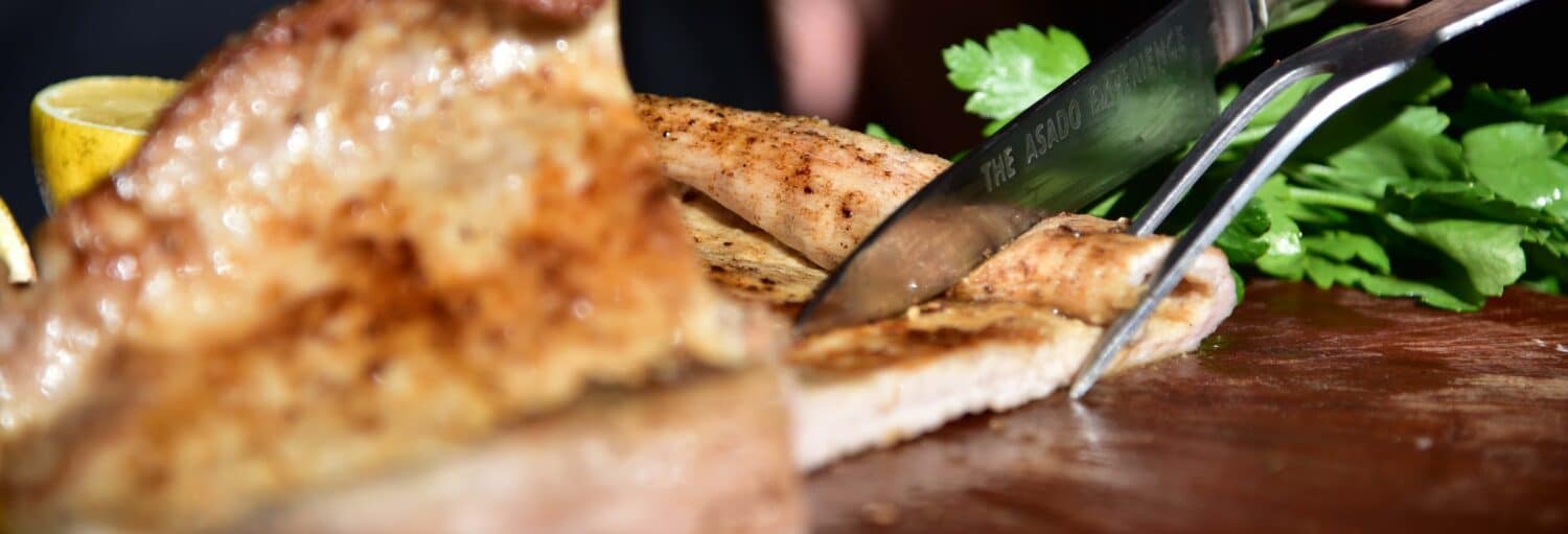 Um close-up de uma faca e uma pinça cortando carne cozida em uma tábua de madeira com ervas frescas por perto. Imagem para ilustrar post sobre oficina de churrasco argentino com degustação em Buenos Aires.