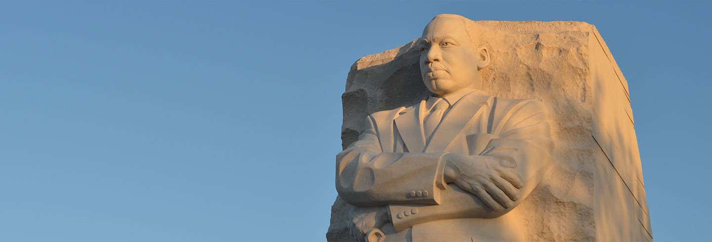 Estátua de pedra de um homem de terno com braços cruzados, em contraste com um céu azul claro.