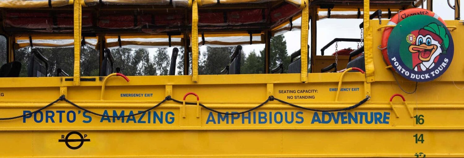 Vista lateral de um ônibus de turismo anfíbio amarelo brilhante com a etiqueta "Porto's Amazing Amphibious Adventure" e o logotipo de um pato de desenho animado. A foto é capa do post de Tour de ônibus anfíbio pelo Porto.