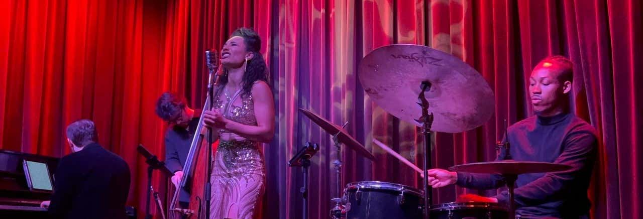 Uma banda de jazz se apresenta no palco com um pianista, um baixista, um cantor em um vestido dourado e um baterista em frente a cortinas vermelhas.