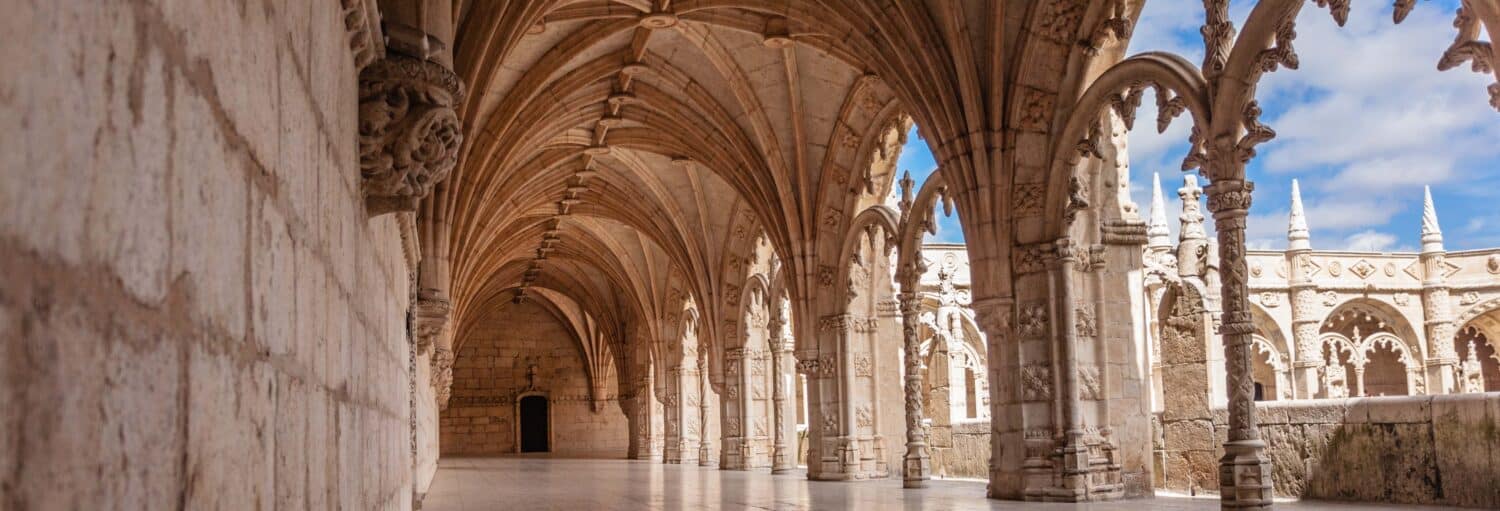 Claustro abobadado em pedra com tetos e colunas arqueadas, com a luz do sol projetando sombras no chão e céu azul visível. Imagem para ilustrar post sobre tour por Belém e mosteiro dos Jerónimos.