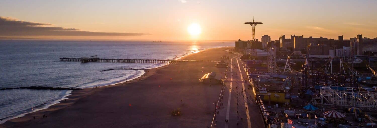 Vista aérea da praia de Coney Island e calçadão ao pôr do sol, com um píer e o horizonte da cidade ao fundo. A foto é capa do post de Tour pelos bairros de Nova York e Coney Island.