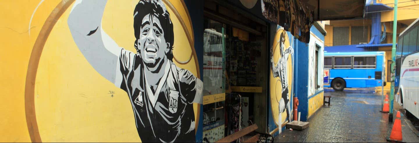 Um mural de um jogador de futebol decora uma parede amarela em uma rua chuvosa com um ônibus azul e cones laranja nas proximidades. Imagem para ilustrar post sobre tour do Maradona por Buenos Aires.