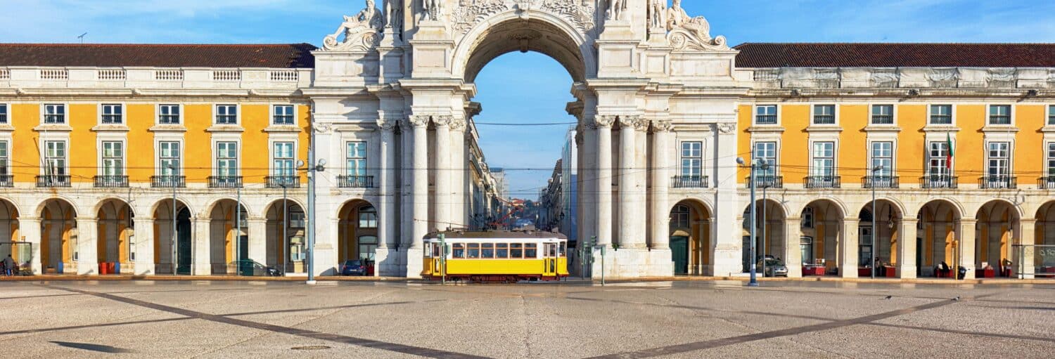 Um bonde amarelo passa em frente ao Arco da Rua Augusta, em uma grande praça pública com prédios amarelos. Imagem para ilustrar post sobre escala em Lisboa? Tour privado saindo do aeroporto.