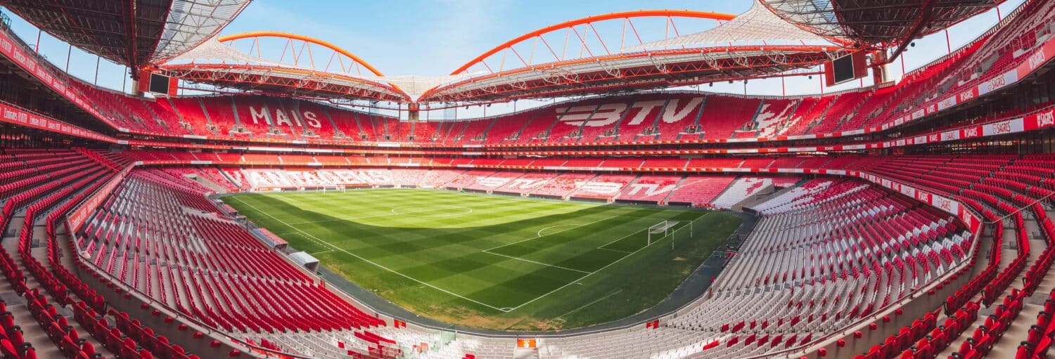 Vista ampla de um estádio de futebol vazio com assentos vermelhos, campo verde e grandes estruturas de telhado arqueadas. Imagem para ilustrar post sobre tour pelo Estádio do Benfica