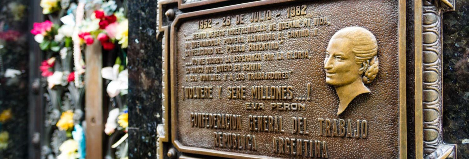 Close-up de uma placa de bronze com o perfil de uma mulher e texto em espanhol; as flores são visíveis no fundo desfocado. Imagem para ilustrar post sobre tour da Eva Perón por Buenos Aires.