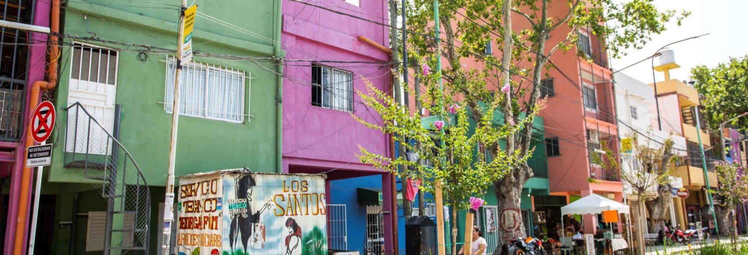 Uma fileira de prédios coloridos, árvores e veículos estacionados se alinham em uma rua em um dia ensolarado. Imagem para ilustrar post sobre tour gastronômico pelo bairro Mugica.
